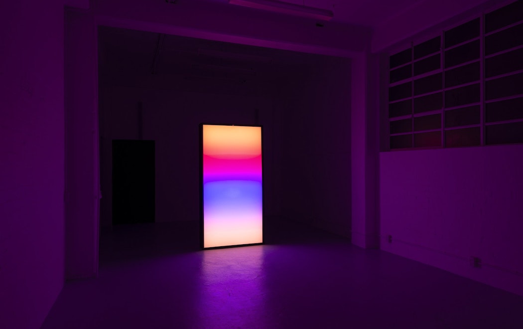 A bright screen illumanates a darkened gallery space. Gradient hues of orange, red, pink, blue bleed against one another slightly